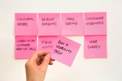 A person holding a pink sticky note that says “Run a usability test”.
