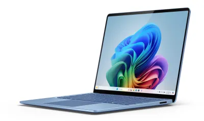 A silver Microsoft Surface laptop. It is open, facing the viewer so its keyboard and mouse are visible, as well as a colorful screen with an abstract, prismatic swirl of paper-thin organic shapes. The Windows 11 taskbar is also visible.