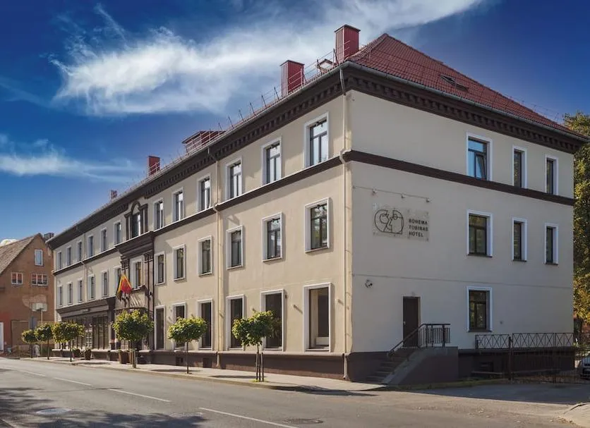 Trijų aukštų viešbučio pastatas su raudonu stogu ir keliais mažais medeliais šaligatvyje prie gatvės.